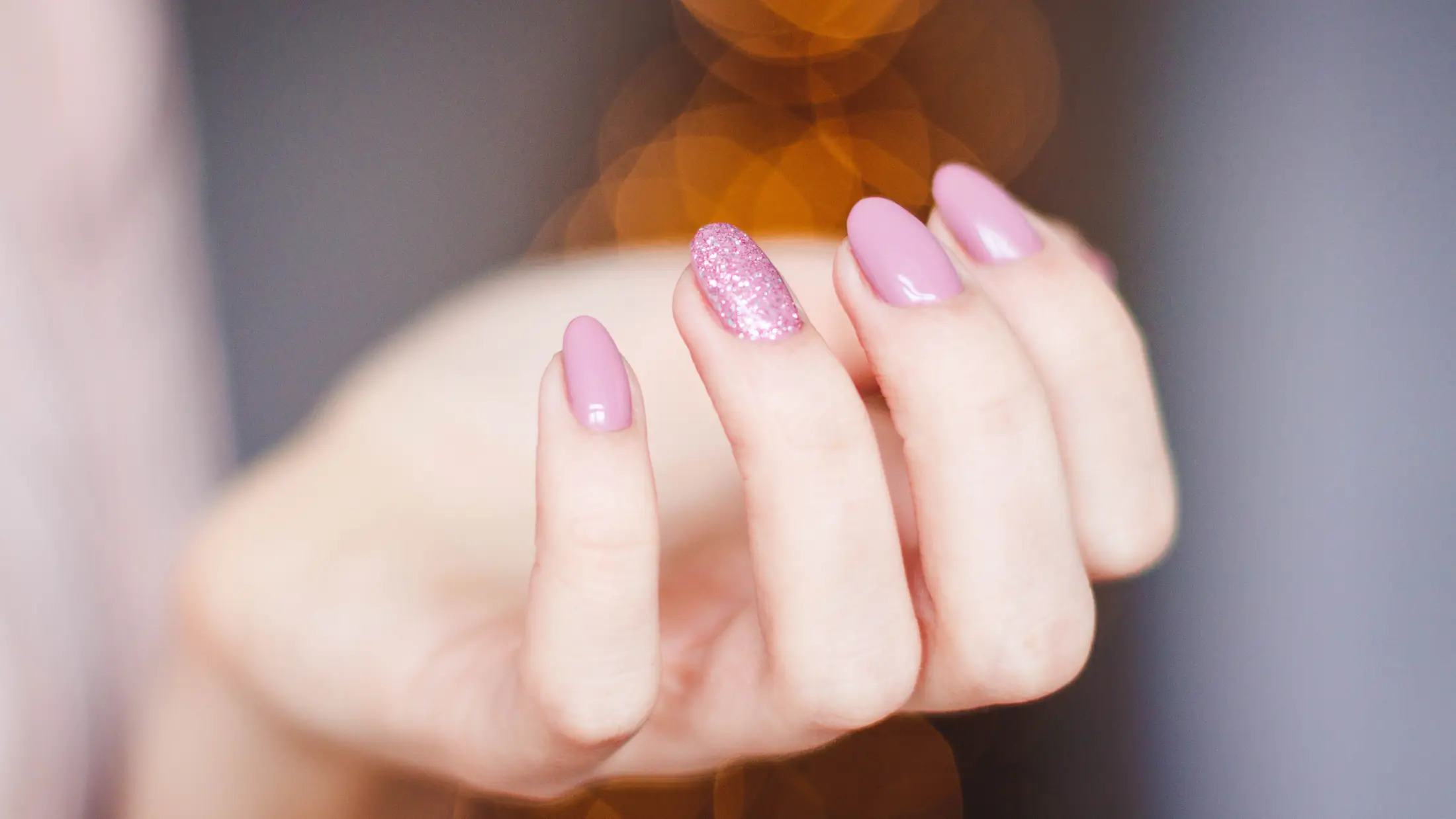 Main aux ongles en amande, pose rose nacrée, lumières dorées en arrière-plan
